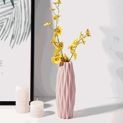 Pink diamond pattern faux ceramic vase with yellow flowers on white surface near candles and framed artwork